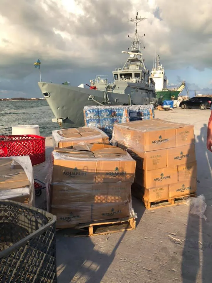 Pallets of bottled water and gallon jugs wrapped in plastic on a dock beside a gray patrol ship flying the flag of the Bahamas.