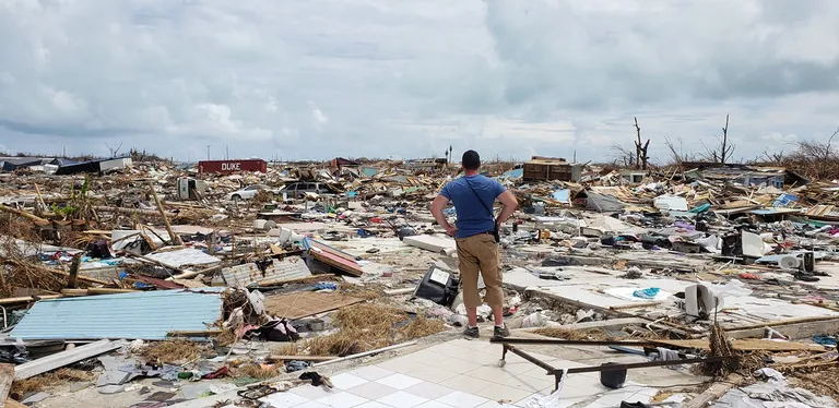 A volunteer in a blue shirt stands on a tiled slab and looks out over a field of flattened homes and debris under a gray sky.