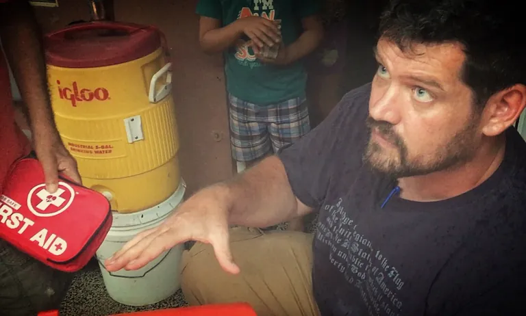 A volunteer kneels beside a yellow water cooler and explains something while a red first aid kit is handed over.