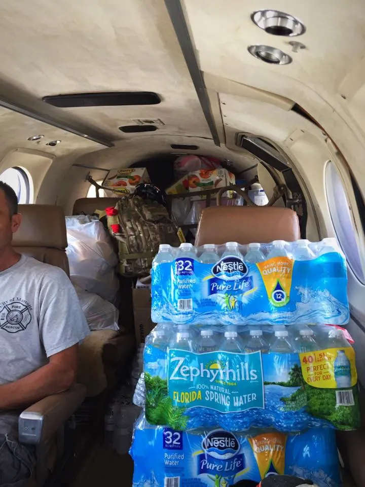 The cabin of a small plane packed with cases of bottled water, bags and boxes, with a passenger in a fire rescue T shirt in the front seat.