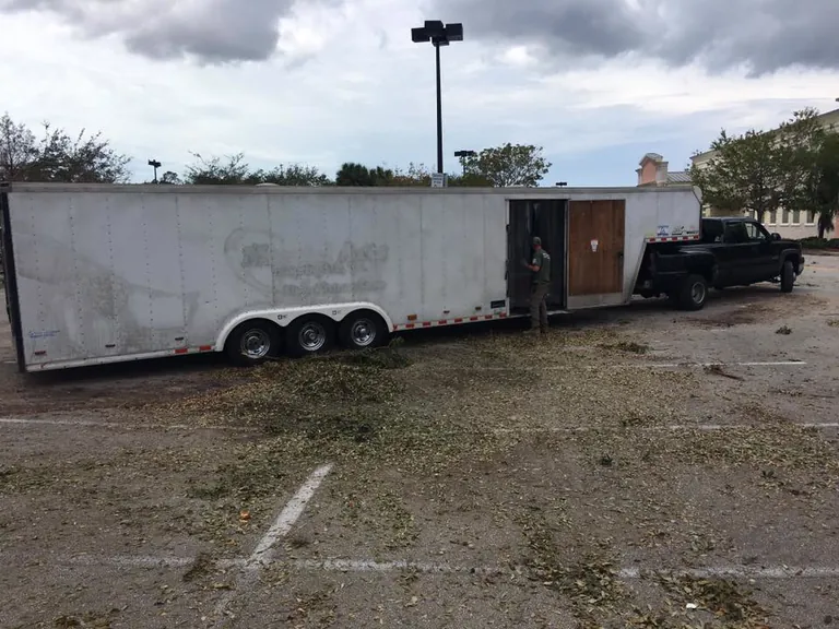 A long white enclosed trailer hitched to a black pickup in a parking lot littered with storm torn leaves.